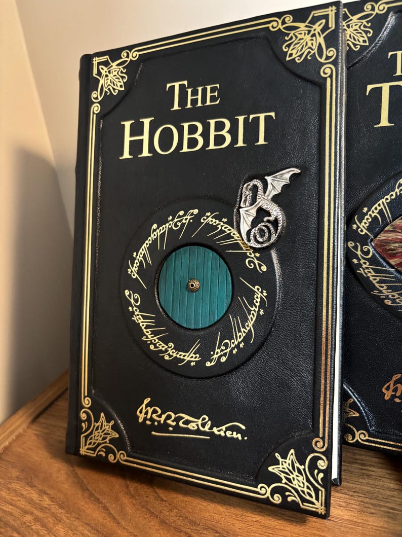 Op de afbeelding: Een zwart leren boek met de titel "The Hobbit" met gouden letters en accenten. De omslag heeft een cirkelvormig ontwerp met een groene deur, een zilveren draak en inscripties. Het boek wordt gepresenteerd op een houten oppervlak.