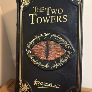 Op de afbeelding: Een zwart boek getiteld "The Two Towers" met gouden letters en decoratieve accenten. De omslag heeft een gedetailleerd oogontwerp in rood, oranje en geel, omringd door schrift. Het boek wordt gepresenteerd op een houten oppervlak.