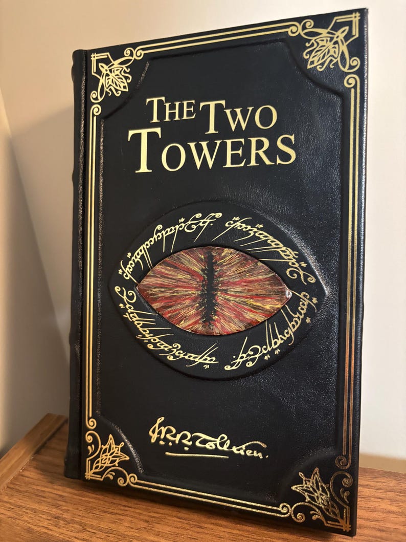 Op de afbeelding: Een zwart boek getiteld "The Two Towers" met gouden letters en decoratieve accenten. De omslag heeft een gedetailleerd oogontwerp in rood, oranje en geel, omringd door schrift. Het boek wordt gepresenteerd op een houten oppervlak.