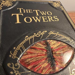 Op de afbeelding: Close-up van een zwarte boekomslag met gouden letters met de tekst "The Two Towers". De omslag heeft een gedetailleerd oogontwerp met rode, gouden en zwarte tinten. Decoratieve gouden filigraan omringt het oog en de titel van het boek.
