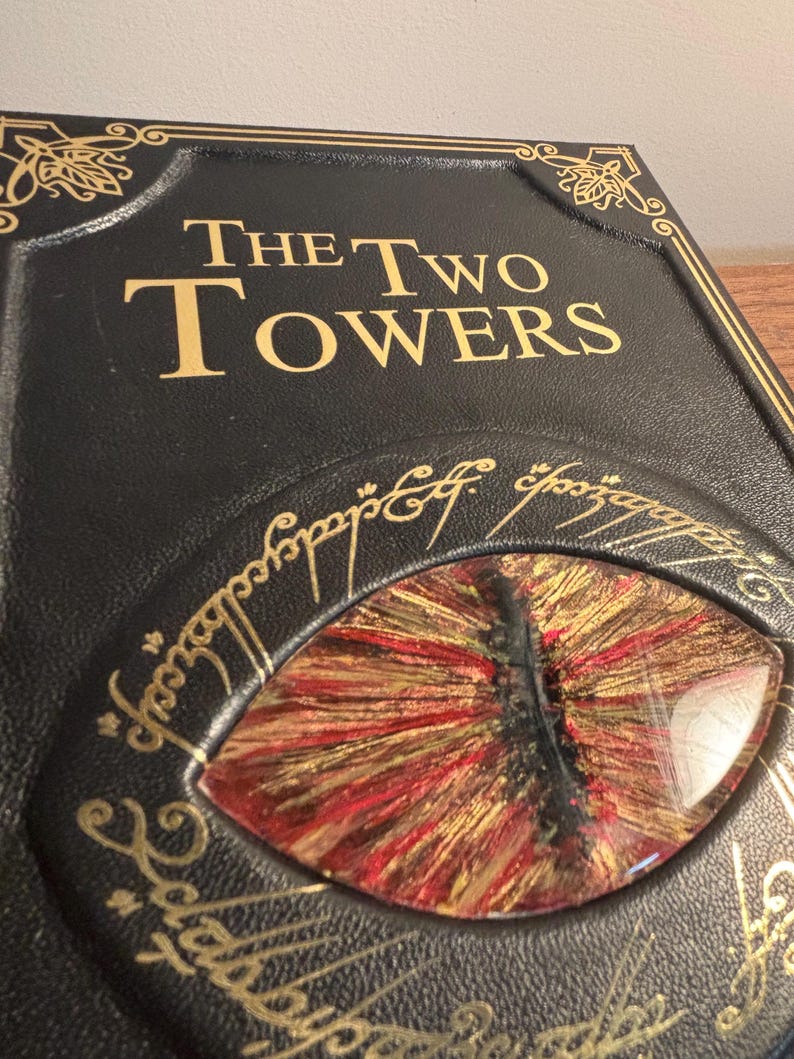 Op de afbeelding: Close-up van een zwarte boekomslag met gouden letters met de tekst "The Two Towers". De omslag heeft een gedetailleerd oogontwerp met rode, gouden en zwarte tinten. Decoratieve gouden filigraan omringt het oog en de titel van het boek.
