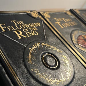 Op de afbeelding: Close-up van een set zwart ledergebonden boeken met gouden letters. Het eerste boek luidt "The Fellowship of the Ring". Een gouden ring aan een ketting staat centraal.