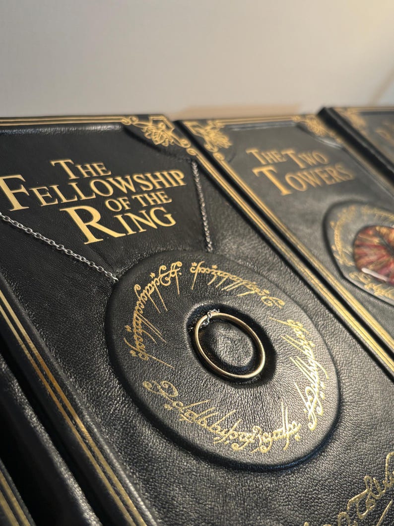 Op de afbeelding: Close-up van een set zwart ledergebonden boeken met gouden letters. Het eerste boek luidt "The Fellowship of the Ring". Een gouden ring aan een ketting staat centraal.
