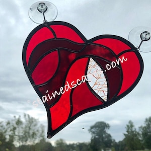 May include: A stained glass heart in shades of red and burgundy, with a black border. The heart-shaped suncatcher has a white and gold patterned section. The text "stainedscar.com" is visible on the glass. Two suction cups are attached for hanging.