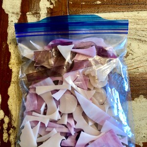 Puede incluir: Una bolsa de pl&aacute;stico transparente llena de trozos de vidrio de forma irregular en tonos blancos, lavanda y morados. La bolsa tiene un cierre de cremallera azul y est&aacute; sobre una superficie de madera envejecida.