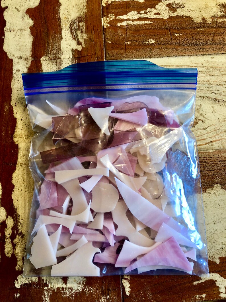 Puede incluir: Una bolsa de pl&aacute;stico transparente llena de trozos de vidrio de forma irregular en tonos blancos, lavanda y morados. La bolsa tiene un cierre de cremallera azul y est&aacute; sobre una superficie de madera envejecida.