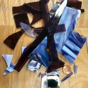 Puede incluir: Vista cenital de una colecci&oacute;n de piezas de vidrio de forma irregular en tonos morados, azules y plateados. Las piezas est&aacute;n dispersas sobre una superficie de madera, con una moneda para dar escala. El vidrio parece ser para un proyecto de mosaico.