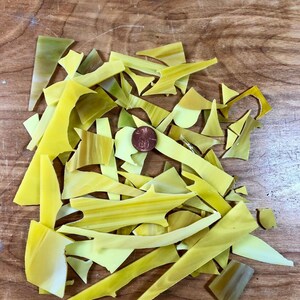 以下が含まれることがあります： さまざまな形や大きさの半透明な黄色のステンドグラスの破片が散らばっています。山の中央付近には銅色の硬貨が1枚置かれており、ガラス片の大きさを比較するためのスケールとして機能しています。