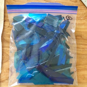 Puede incluir: Una bolsa de pl&aacute;stico transparente llena de trozos de vidrio te&ntilde;ido azul de varios tama&ntilde;os. La bolsa est&aacute; sellada con un cierre de cremallera azul. La bolsa est&aacute; etiquetada con el n&uacute;mero "1 lb" en rotulador negro.
