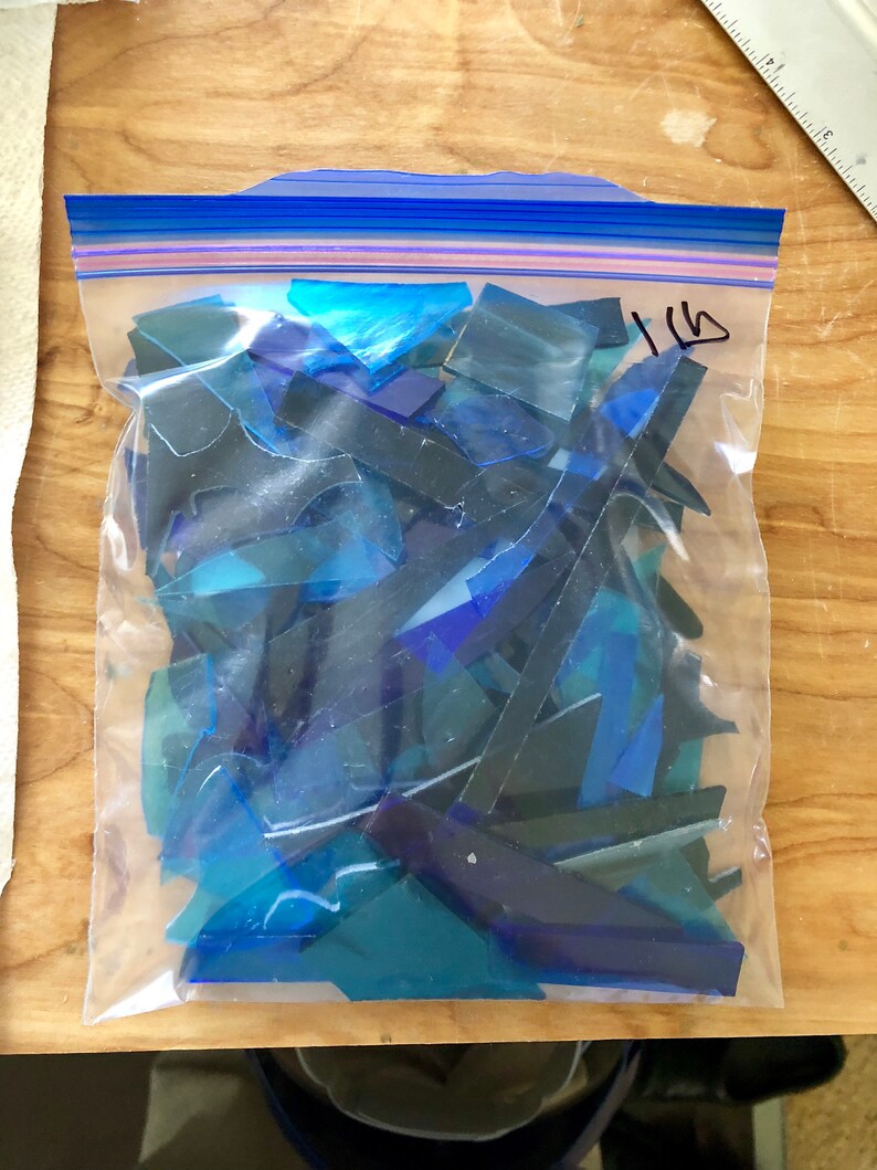 Puede incluir: Una bolsa de pl&aacute;stico transparente llena de trozos de vidrio te&ntilde;ido azul de varios tama&ntilde;os. La bolsa est&aacute; sellada con un cierre de cremallera azul. La bolsa est&aacute; etiquetada con el n&uacute;mero "1 lb" en rotulador negro.