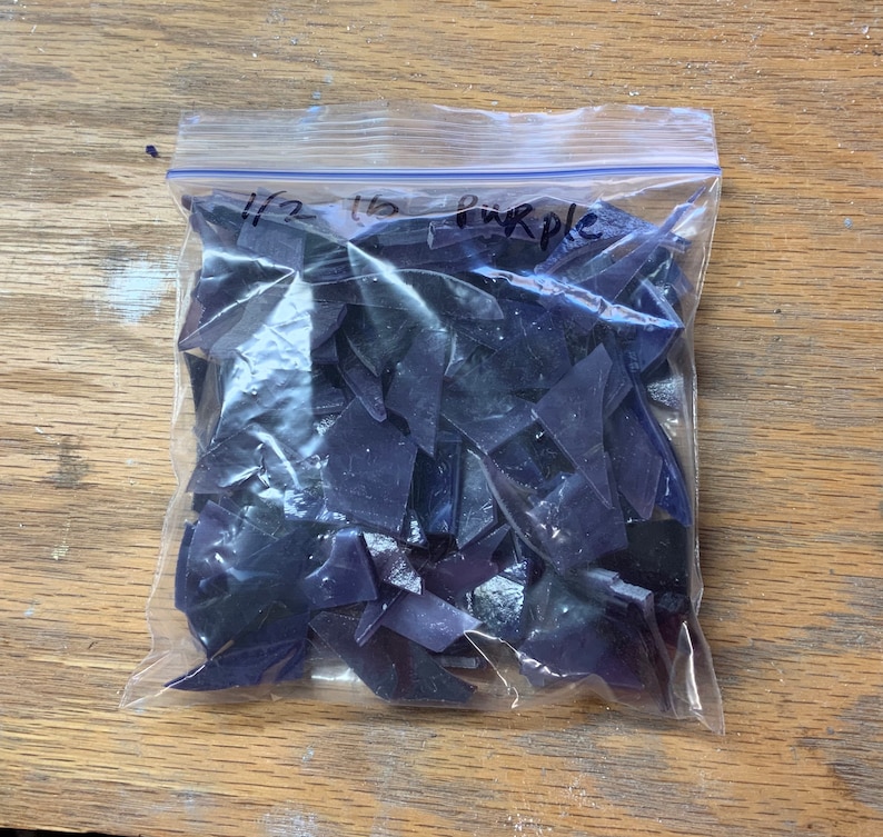 以下が含まれることがあります： 透明な再封可能なプラスチックバッグに、小さく不規則な半透明の紫色の破片が入っています。バッグには黒い手書き文字で「1/2 lb purple」と書かれており、内容物が紫色の破片であることを示しています。