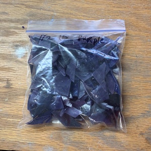 Puede incluir: Una bolsa de pl&aacute;stico transparente llena de trozos de vidrio morado oscuro de forma irregular. La bolsa est&aacute; etiquetada con "1/2 lb Purple" con un rotulador. Las piezas parecen ser para proyectos de mosaicos o vidrieras.