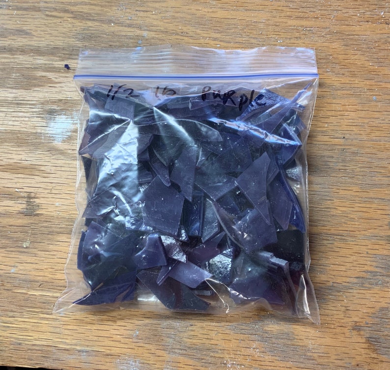 Puede incluir: Una bolsa de pl&aacute;stico transparente llena de trozos de vidrio morado oscuro de forma irregular. La bolsa est&aacute; etiquetada con "1/2 lb Purple" con un rotulador. Las piezas parecen ser para proyectos de mosaicos o vidrieras.