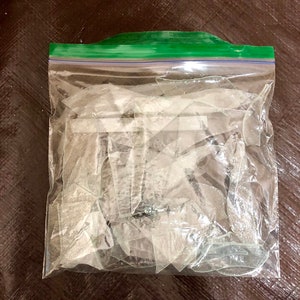 以下が含まれることがあります： 透明な再封可能なプラスチック製の保存袋に、ギザギザした半透明の割れたガラスの破片が多数入っています。袋の上部には特徴的な緑色のプラスチック製ジッパーシールが付いており、鋭利で不規則なガラスの破片をしっかりと中に保持しています。