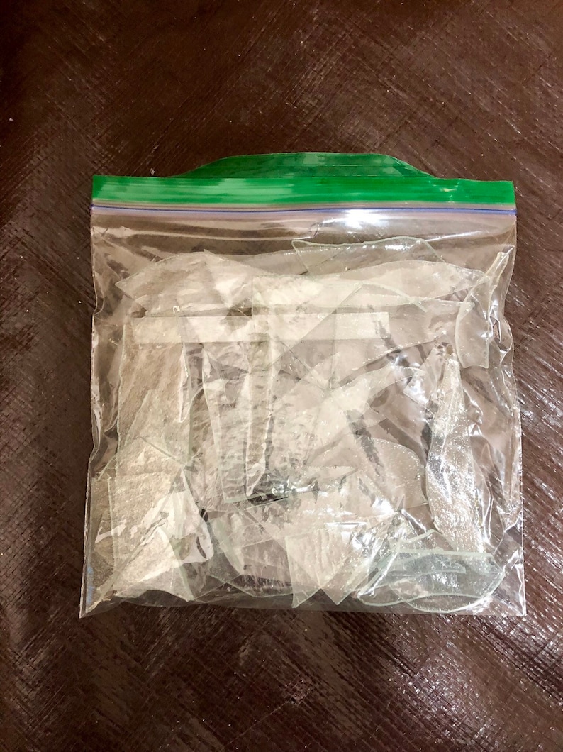 以下が含まれることがあります： 透明な再封可能なプラスチック製の保存袋に、ギザギザした半透明の割れたガラスの破片が多数入っています。袋の上部には特徴的な緑色のプラスチック製ジッパーシールが付いており、鋭利で不規則なガラスの破片をしっかりと中に保持しています。