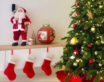May include: A decorated Christmas tree with red and gold ornaments, candy canes, and lights. A Santa Claus figurine stands next to a large, round, red and gold Christmas ornament. Three red stockings with gold lettering hang on a white shelf.