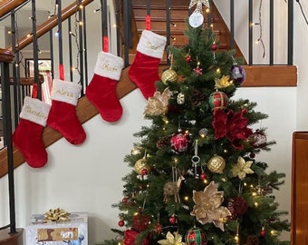 May include: A decorated Christmas tree with red and gold ornaments, pine cones, and red velvet poinsettias. There are four stockings hanging on a railing behind the tree, each with a name embroidered on it: Theodore, Alexa, Mum, and Dad.