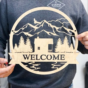 May include: A wooden welcome sign featuring a mountain scene, a cabin, and a lake. The sign is round and has a banner that reads "WELCOME".