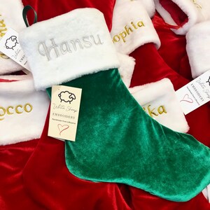May include: A green and red velvet Christmas stocking with white faux fur trim. The stocking is personalized with the name "Hansu" in silver embroidery. A white sheep logo with the text "White Sheep Embroidery Handmade from Lithuania" is attached to the stocking.