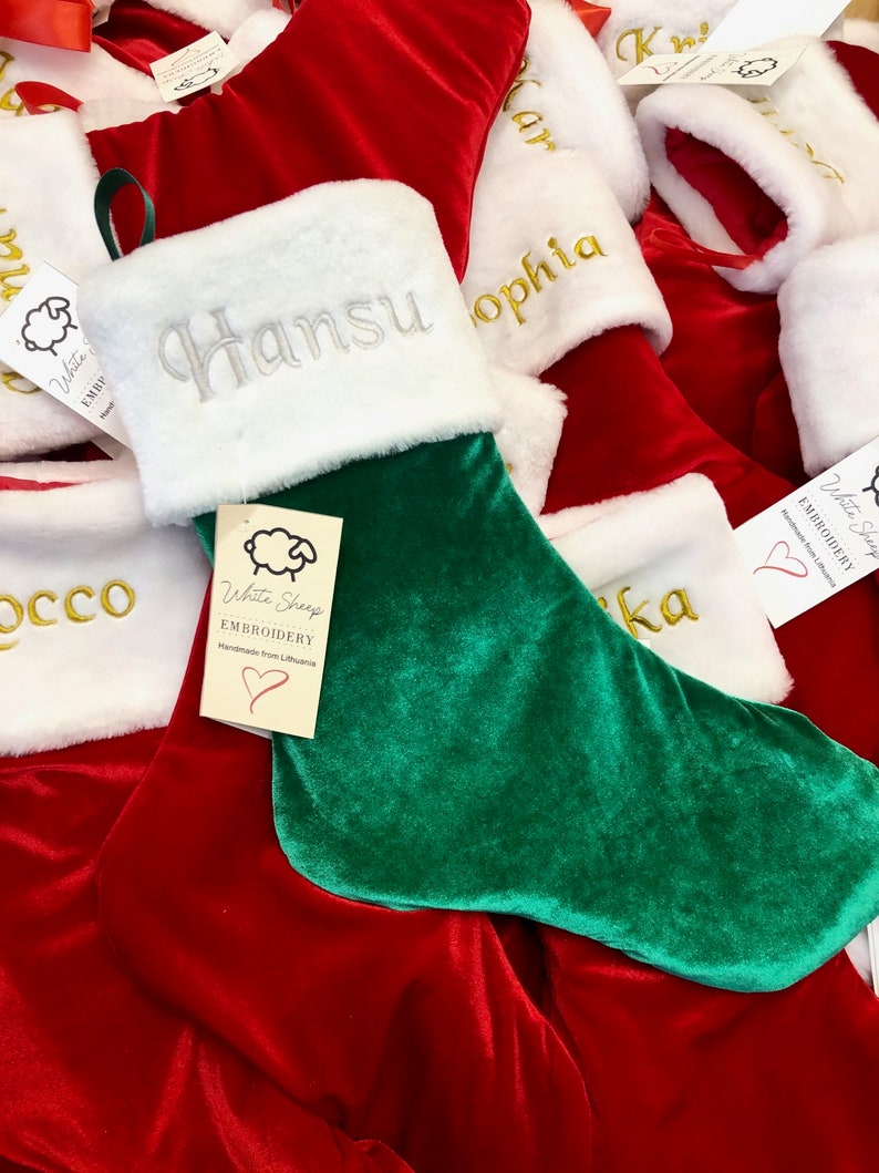 May include: A green and red velvet Christmas stocking with white faux fur trim. The stocking is personalized with the name "Hansu" in silver embroidery. A white sheep logo with the text "White Sheep Embroidery Handmade from Lithuania" is attached to the stocking.