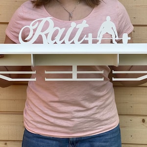Può includere: Un porta medaglie da parete bianco con la parola "Raít" e una silhouette di una persona che solleva pesi. Il porta medaglie ha tre file di ganci per appendere le medaglie.
