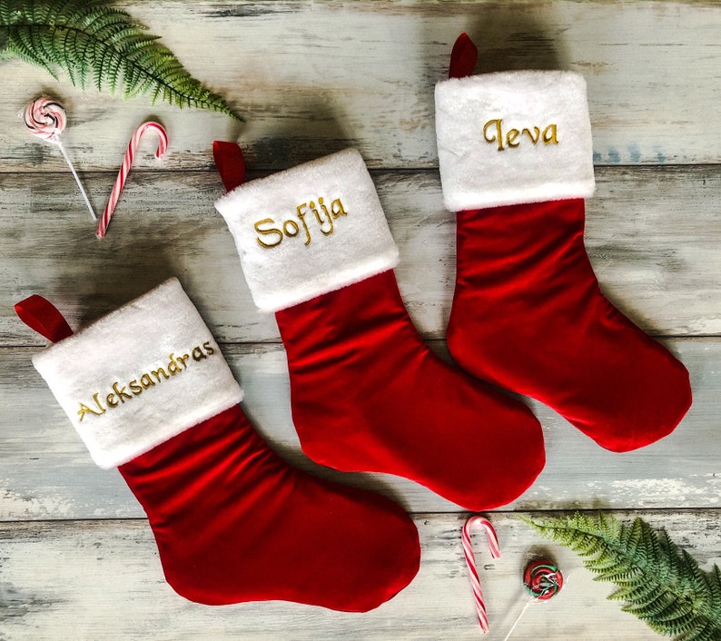 May include: Three red Christmas stockings with white cuffs and gold embroidered names: "Aleksandras", "Sofija", and "Ieva".