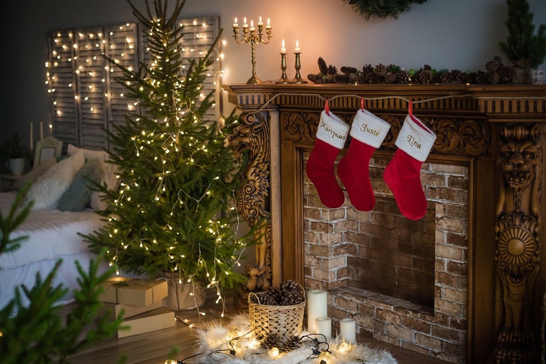 May include: A decorated fireplace mantel with three red stockings hanging from it. The stockings are personalized with the names "Merrymas", "Justie", and "Lina".