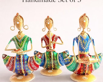 May include: A handmade set of 3 metal figurines featuring stylised figures in vibrant green, red, and blue outfits with intricate painted patterns. Each figure stands on a circular base, with one holding a small plate, one playing a flute, and one playing a drum.