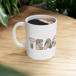 May include: White ceramic mug filled with dark coffee. The word "TEACHER" is printed on the mug in a patchwork quilt design with various colours and patterns. The mug sits on a light wood surface.