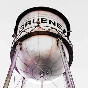 May include: A large, white water tower with the word "GRUENE" in black letters on the side. The tower is made of metal and has a round, bulbous shape. It is supported by three sets of metal legs.