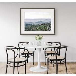 May include: A white round table with a white top and a white base. Four black chairs with woven seats surround the table. A framed photograph of a misty mountain landscape hangs on the wall behind the table.