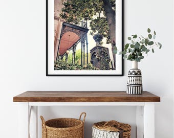 May include: A framed print of a historic building with a pink awning, black ironwork, and lush greenery. The print hangs above a white and brown wooden console table. Two woven baskets sit below, and a vase with greenery is on the table.