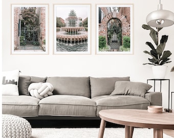 May include: Three framed prints of a brick building with a wrought iron gate, a fountain with a pineapple sculpture, and a brick archway with a wrought iron gate. The prints are hanging on a white wall above a gray sofa.