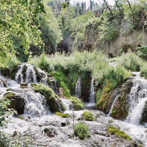 May include: A cascading waterfall flows over moss-covered rocks in a lush green forest setting.