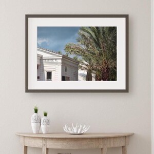 May include: A framed wall art print featuring a white building with the text SEASIDE, FL visible on the facade, positioned next to a large palm tree under a blue sky. Below the frame are two white textured vases and a white coral-shaped bowl.