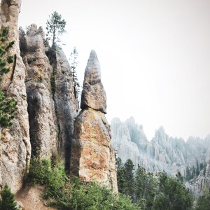 May include: A tall, thin rock formation rises from a rocky landscape, with other rock formations in the background. The rock formation is a light brown color and has a rough texture. The background is a light gray color and is out of focus.