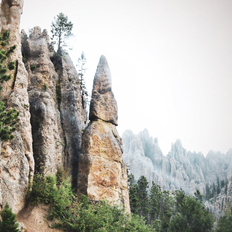 May include: A tall, thin rock formation rises from a rocky landscape, with other rock formations in the background. The rock formation is a light brown color and has a rough texture. The background is a light gray color and is out of focus.