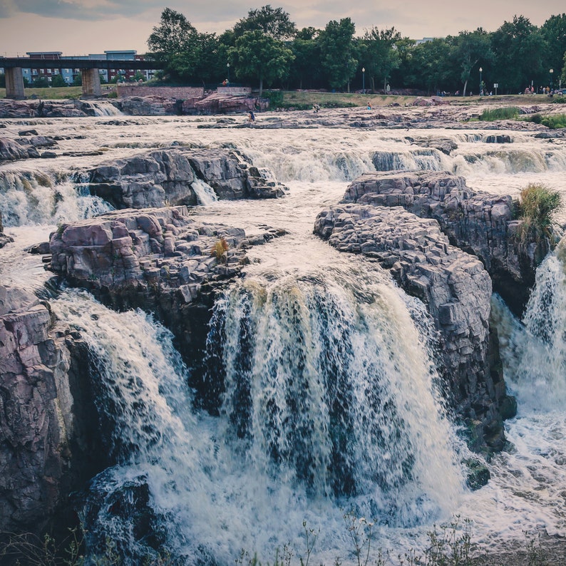 May include: A wide shot of a waterfall cascading over large, gray rocks. The water is white and foamy, and the rocks are covered in moss and lichen. The waterfall is surrounded by trees and greenery.