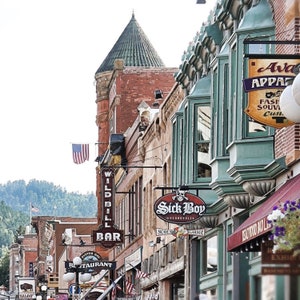 May include: A street scene in a historic town with a variety of shops and businesses. The buildings are made of brick and wood and have a rustic charm. There are signs for "Wild Bill Bar", "Sick Boy Motorcycles", and "Avar Apparel".