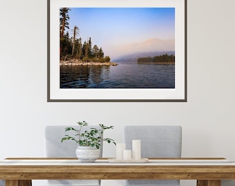 May include: A framed landscape photograph of a calm lake bordered by a rocky shoreline and dense evergreen trees under a hazy blue sky. The scene features soft light reflecting on the water surface with distant mountains obscured by light fog.