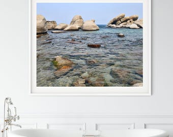 May include: A framed wall art print featuring a coastal scene with large, smooth beige boulders emerging from clear, shallow turquoise water under a pale blue sky. The composition captures a tranquil shoreline with submerged rocks visible.
