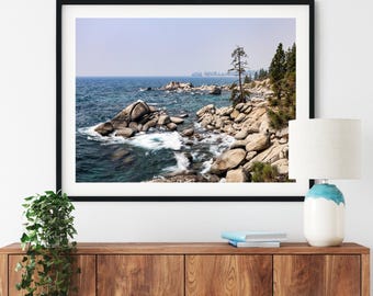 May include: A framed landscape photograph of a rocky shoreline meeting a deep blue sea. Large granite boulders line the coast, with evergreen trees growing along the edge. The water shows white foam where waves break against the stones.