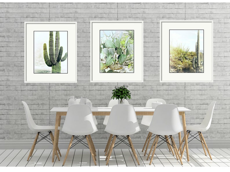 Pu&ograve; includere: Tre acquerelli incorniciati di cactus in un ambiente desertico. I dipinti sono appesi a un muro di mattoni grigi sopra un tavolo da pranzo bianco con sei sedie bianche.