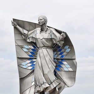 May include: A silver statue of a woman with long braids wearing a flowing dress and a large, geometric patterned cape. The statue is outdoors against a cloudy sky.
