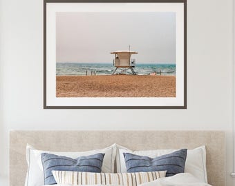 May include: A framed wall art print featuring a white lifeguard tower standing on a sandy beach. The scene captures the ocean horizon under a pale sky, with the structure elevated on metal stilts. The frame is dark brown and rectangular.