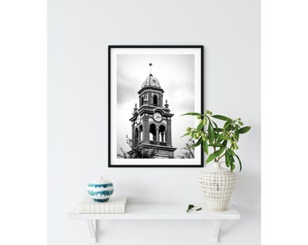 Puede incluir: Fotografía en blanco y negro de una alta torre del reloj con una esfera del reloj y una aguja. La torre está hecha de ladrillo y tiene un campanario. La foto está enmarcada en un marco negro.