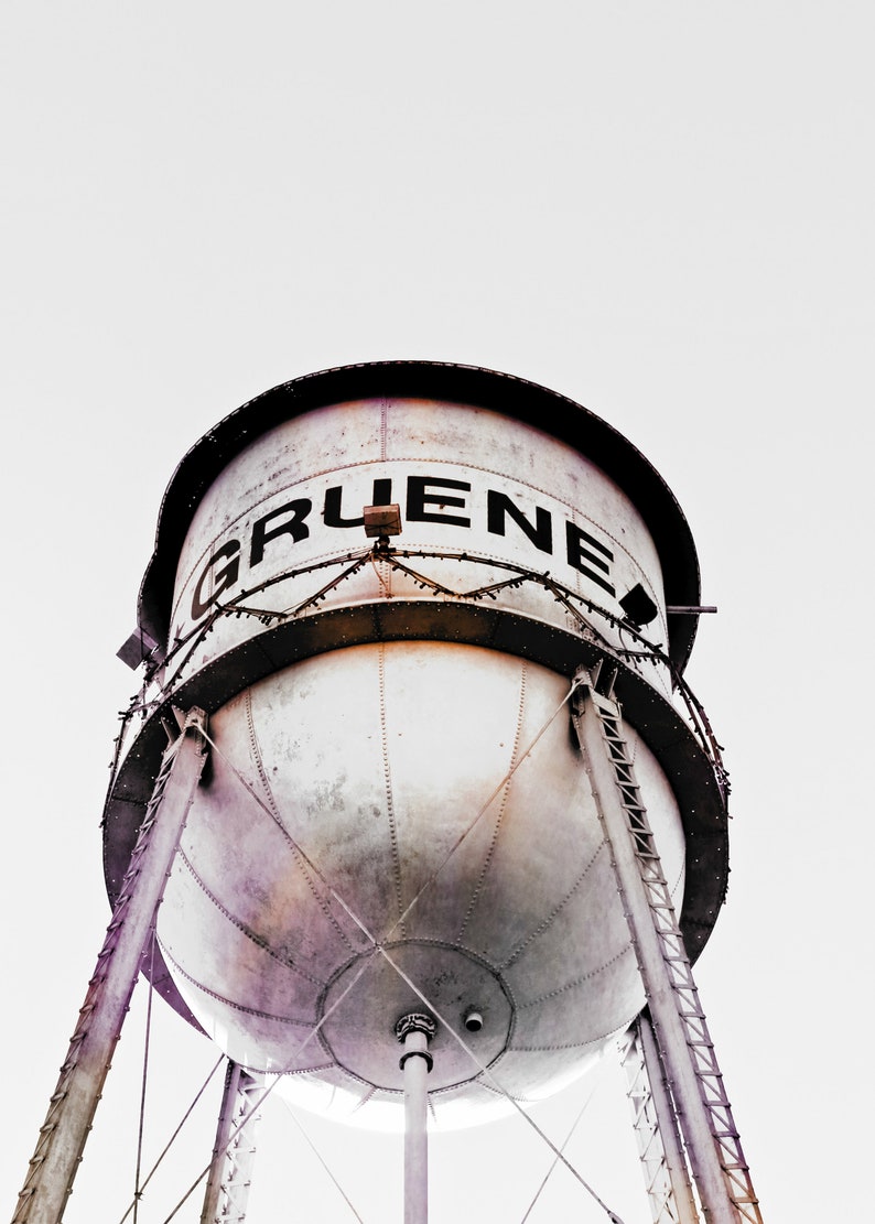 May include: A large, white water tower with the word "GRUENE" in black letters on the side. The tower is made of metal and has a round, bulbous shape. It is supported by three sets of metal legs.