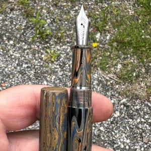 May include: A fountain pen with a silver nib and a patterned barrel in shades of brown, blue, and black. The pen is partially disassembled, with the cap removed, revealing the intricate design and metal components.