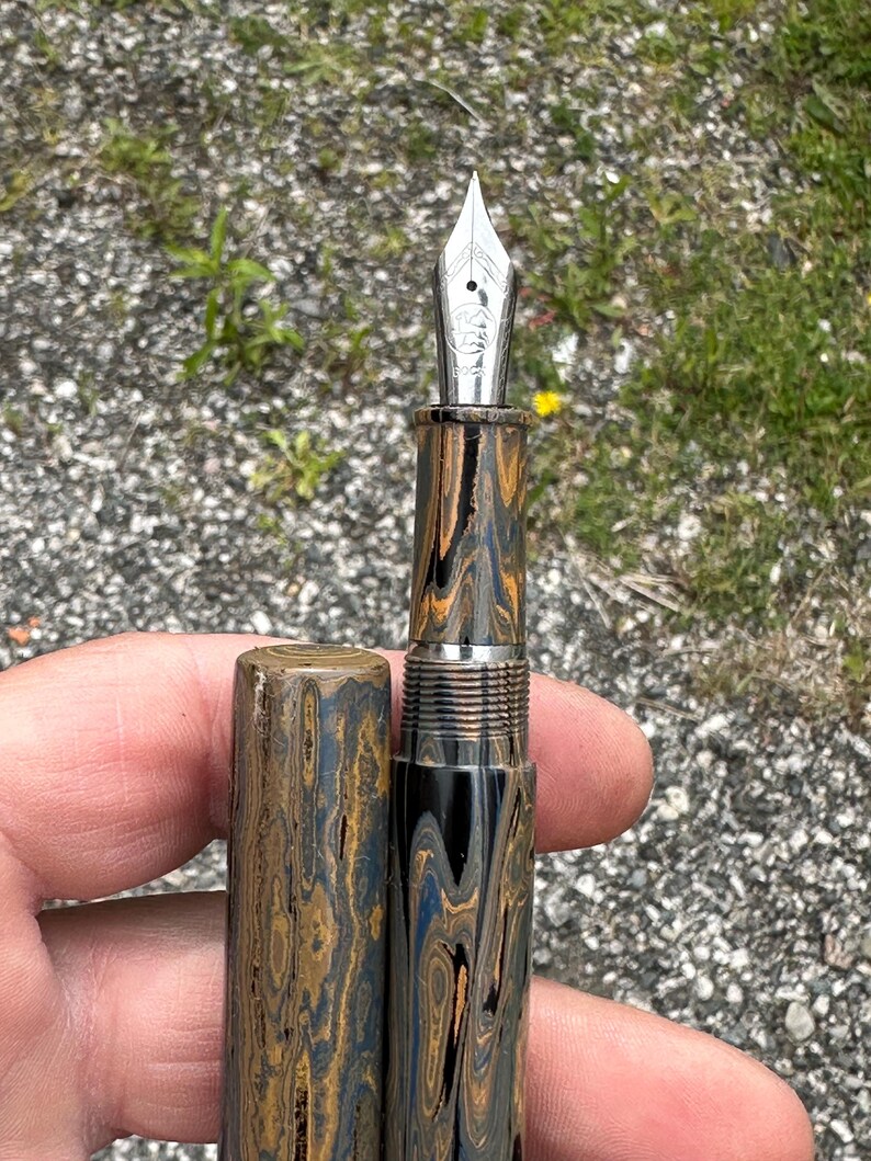 May include: A fountain pen with a silver nib and a patterned barrel in shades of brown, blue, and black. The pen is partially disassembled, with the cap removed, revealing the intricate design and metal components.