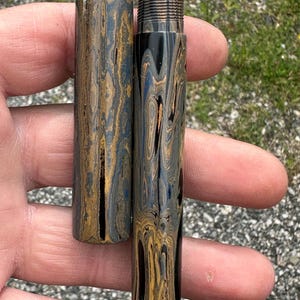 May include: A handcrafted pen and pen case with a unique swirling pattern in shades of black, gold, and blue. The pen features a silver metal band near the nib. The pen case is cylindrical and matches the pen's design.